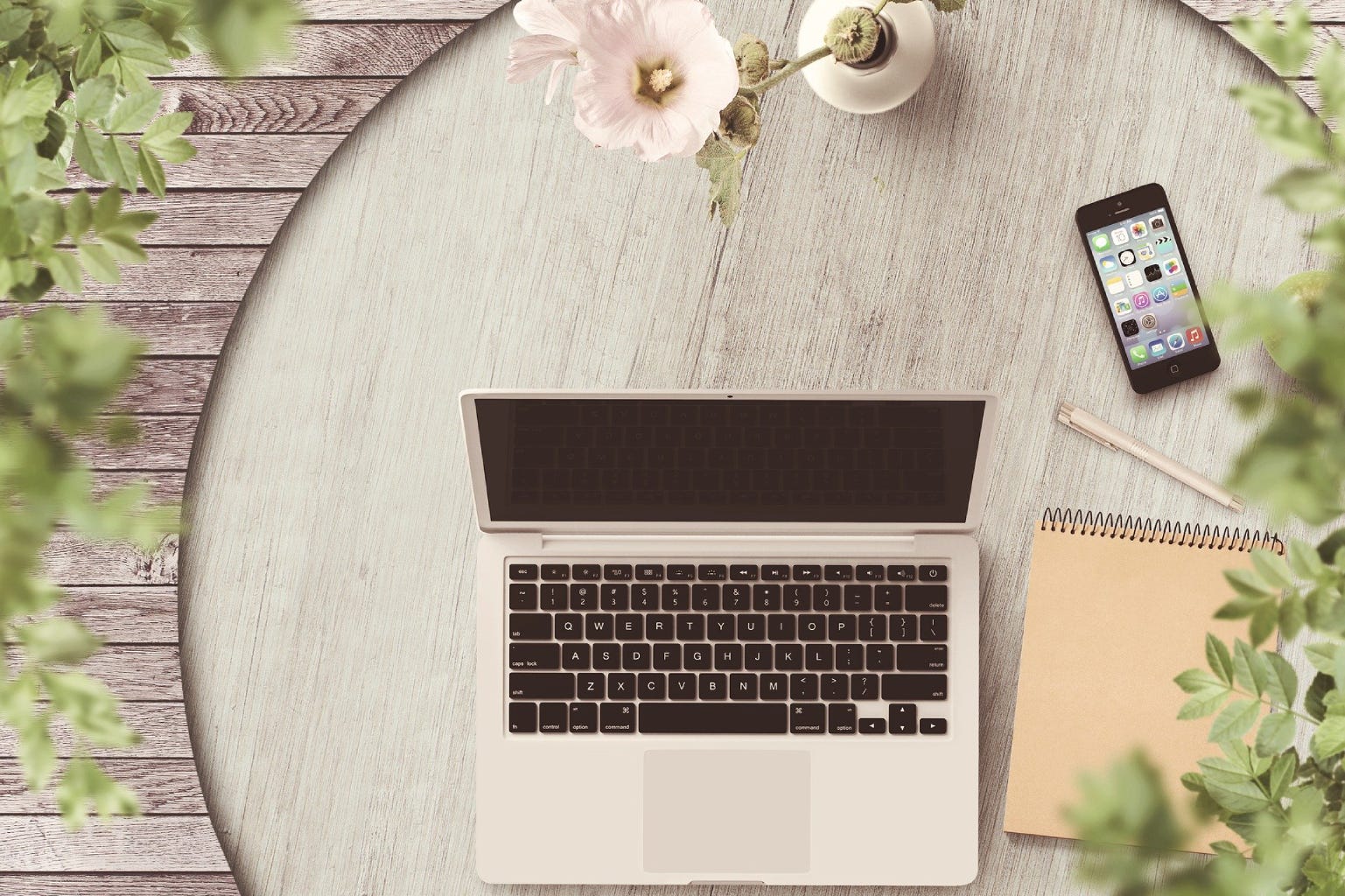 Laptop and iphone on a table outside with green vegetation around.