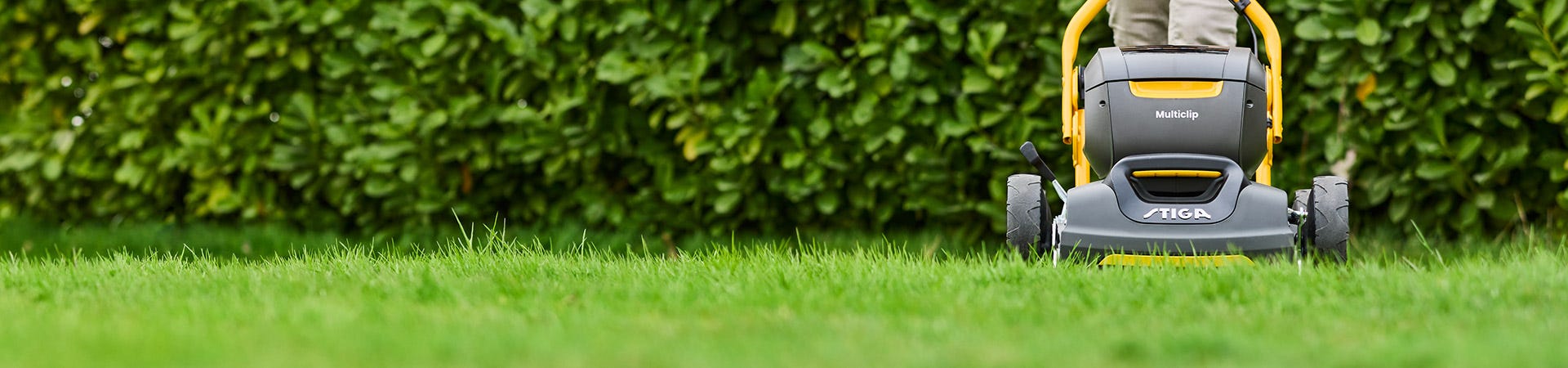 Woman cutting grass along a hedge with a STIGA Multiclip mulching lawn mower with the STIGA Fulcrum handlebar
