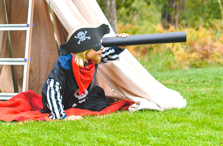 Un enfant blond à l'extérieur d'une tente dans le jardin, habillé en pirate et utilisant le tube d'un souffleur de feuilles comme télescope.