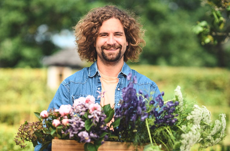Junger Mann mit braunen Haaren und Bart hält eine Holzkiste voller Blumen vor einem grünen Hintergrund